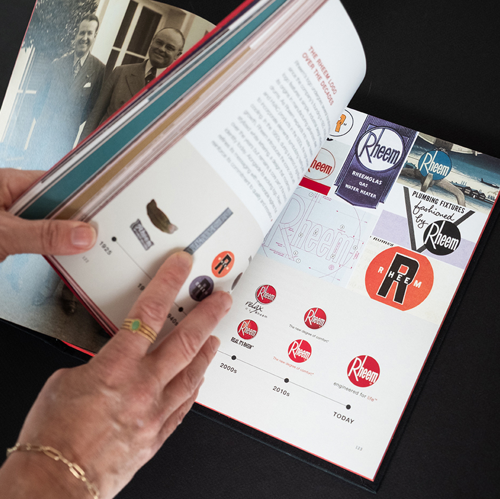 Hands turning the pages of a custom-designed corporate history book for Rheem, showcasing a beautifully curated visual timeline of vintage brand logos and corporate identity evolution.